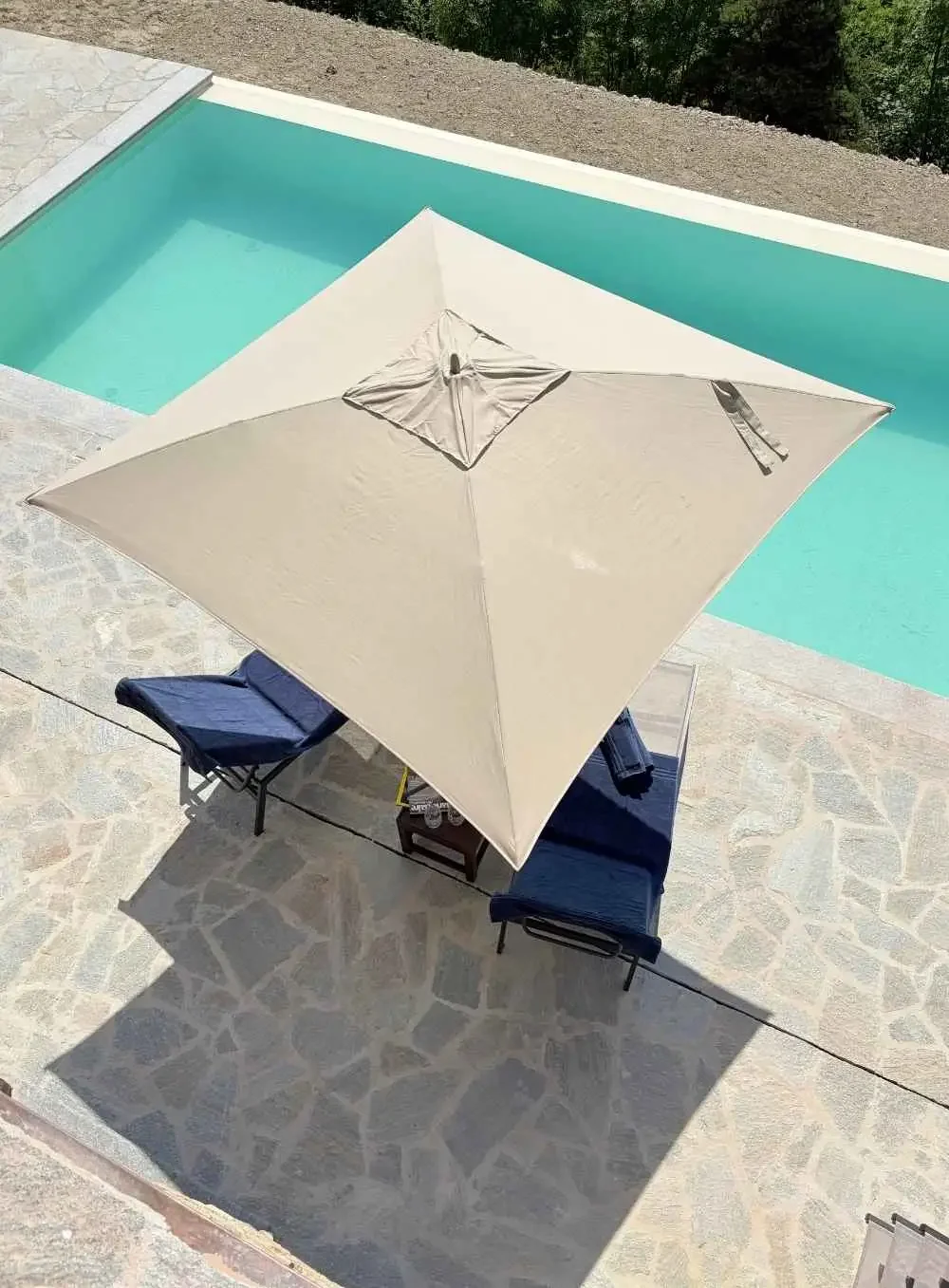 A swimming pool with a beige umbrella providing shade to a patio area with two lounge chairs and a small table. The pool has a stone deck surrounding it.