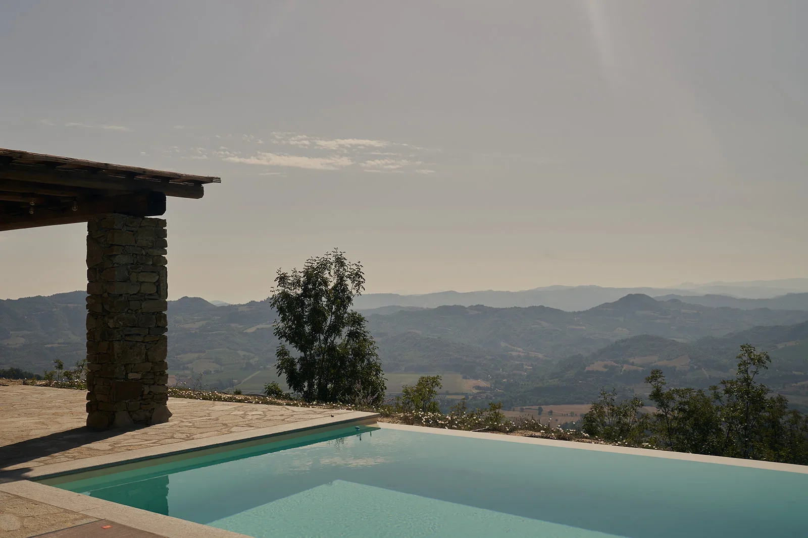 Scenic view of a mountain landscape with a clear sky, featuring part of a stone patio, a swimming pool, and some trees in the foreground.