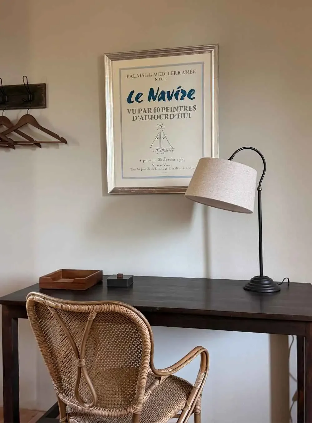 A home office desk with a rattan chair, a black table with a tray and a box, a framed poster with French text about a sailing theme, a floor lamp, and a wall hook with wooden hangers in a neutral-colored room.