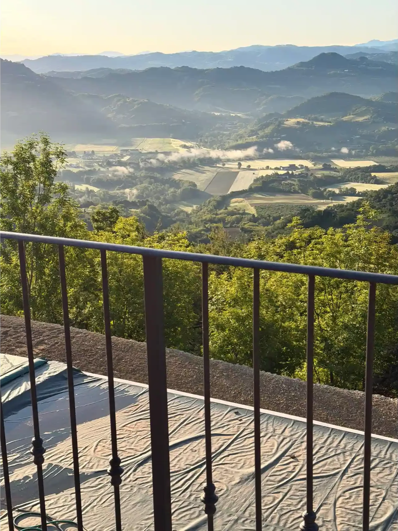 View of a scenic valley with rolling hills and mountains in the background, seen from a balcony with a metal railing, and a blanket or mat spread on the ground in the foreground.