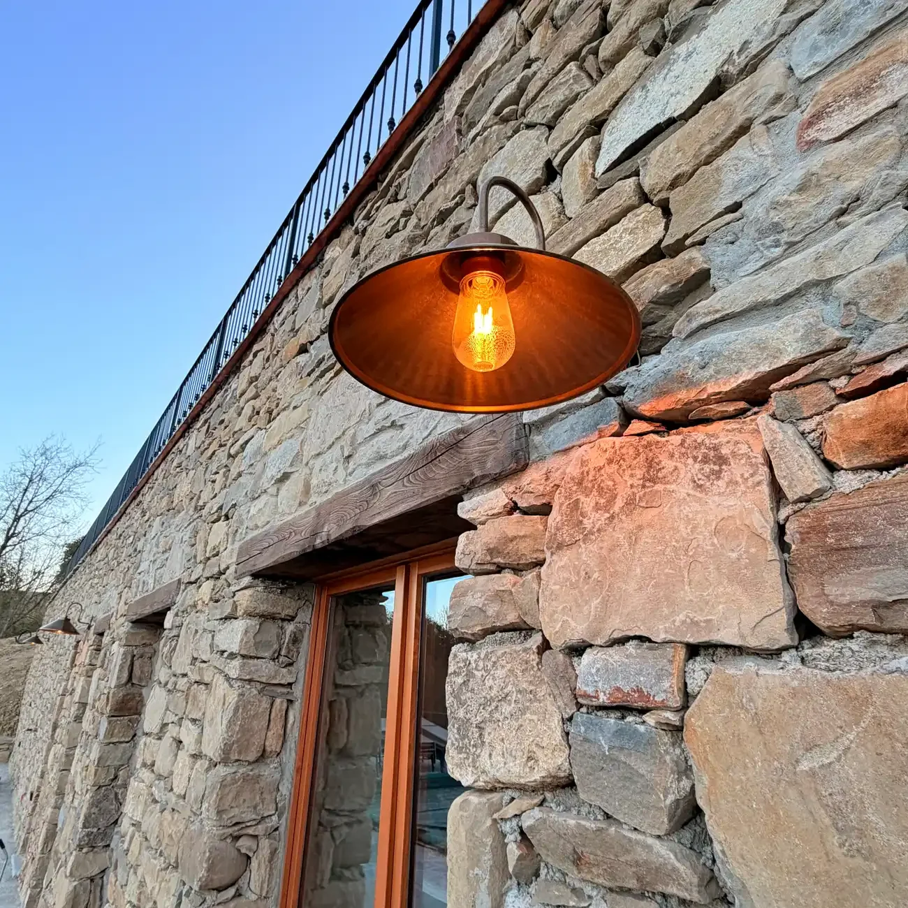 Close-up of a rustic exterior stone wall on a building with a window and outdoor wall-mounted lamp with an orange glow, under a clear blue sky.