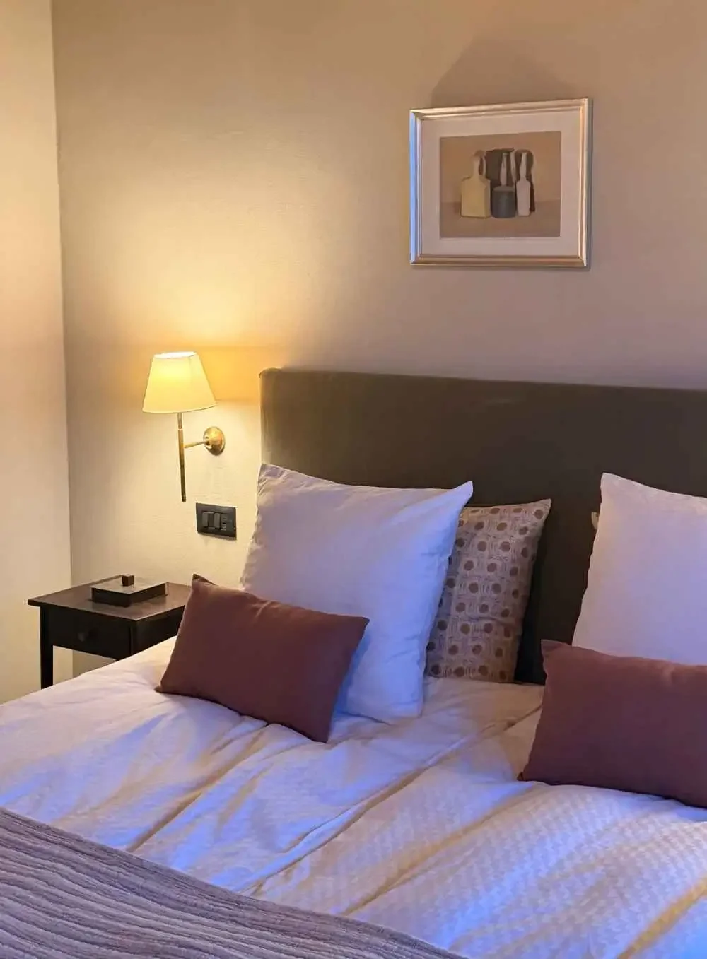 A cozy hotel bedroom with a neatly made bed, multiple pillows, wall-mounted lamp, a small side table, and framed artwork depicting bottles on a beige wall.