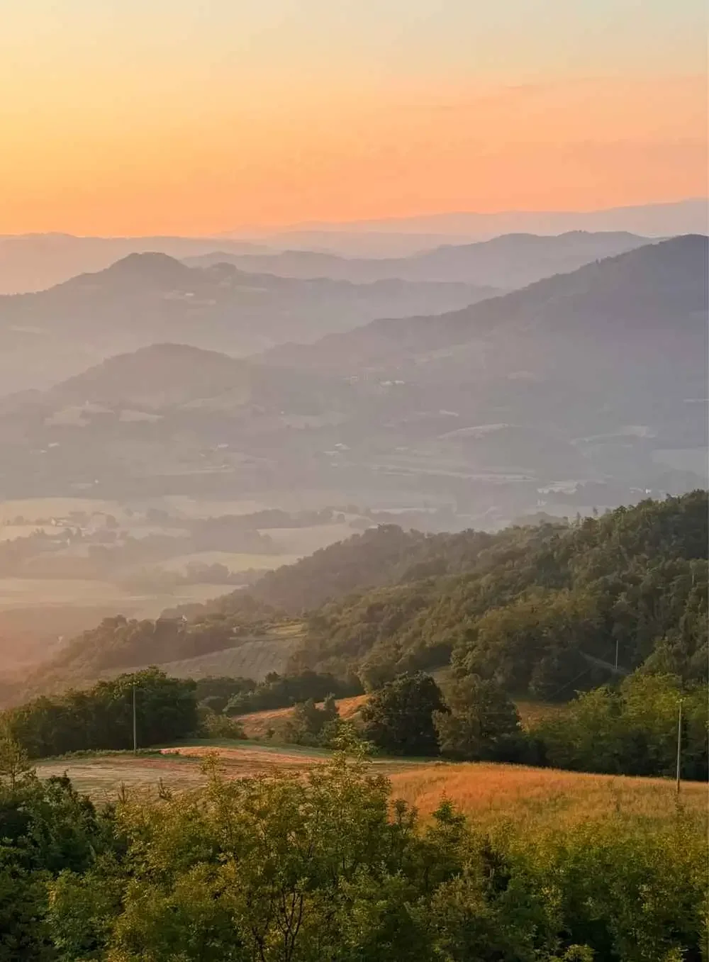 A landscape of layered hills and mountains at sunset, with a mix of green forests and open fields.