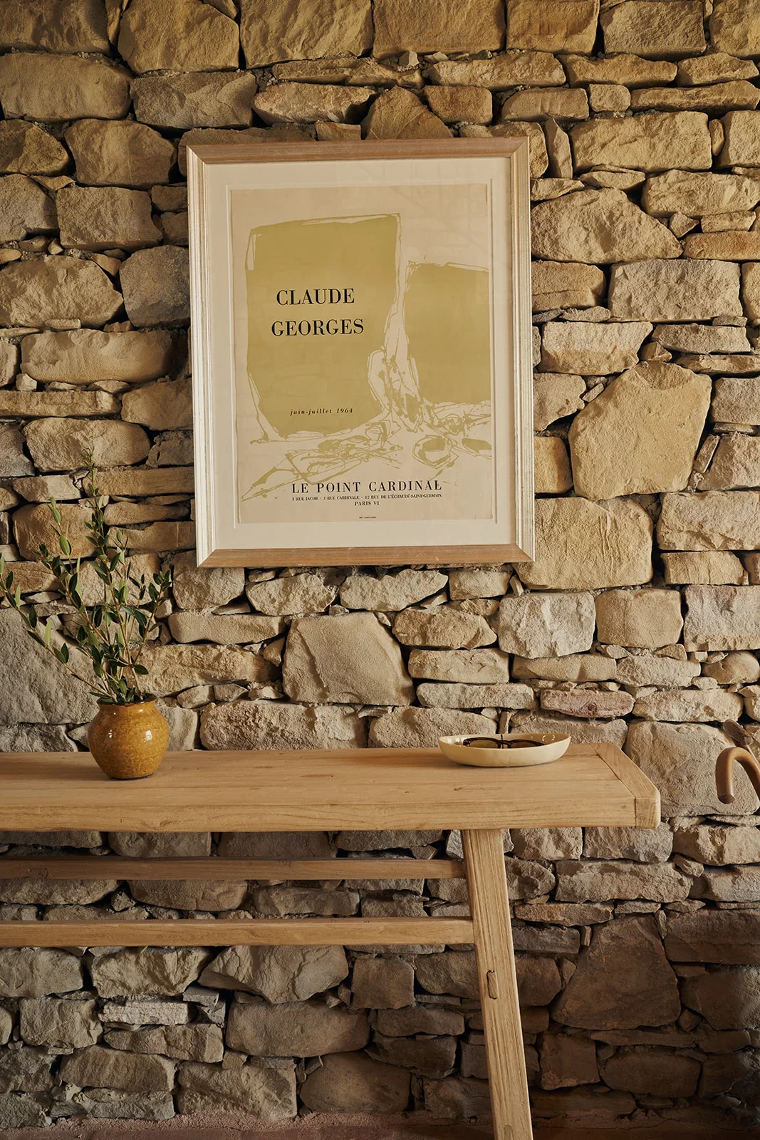 Decorative interior wall with framed poster of Claude Georges, a stone wall, a wooden table with a small pottery vase holding greenery, and a ceramic dish with utensils.
