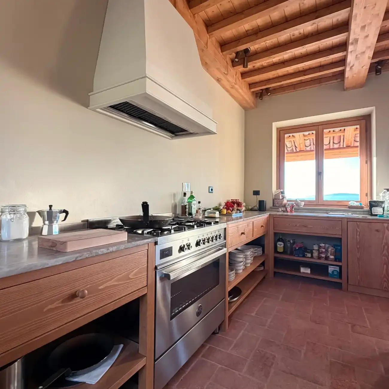 A rustic kitchen with wooden cabinets, a stainless steel oven, and a window with a view outside. The countertop holds various kitchen items and ingredients.