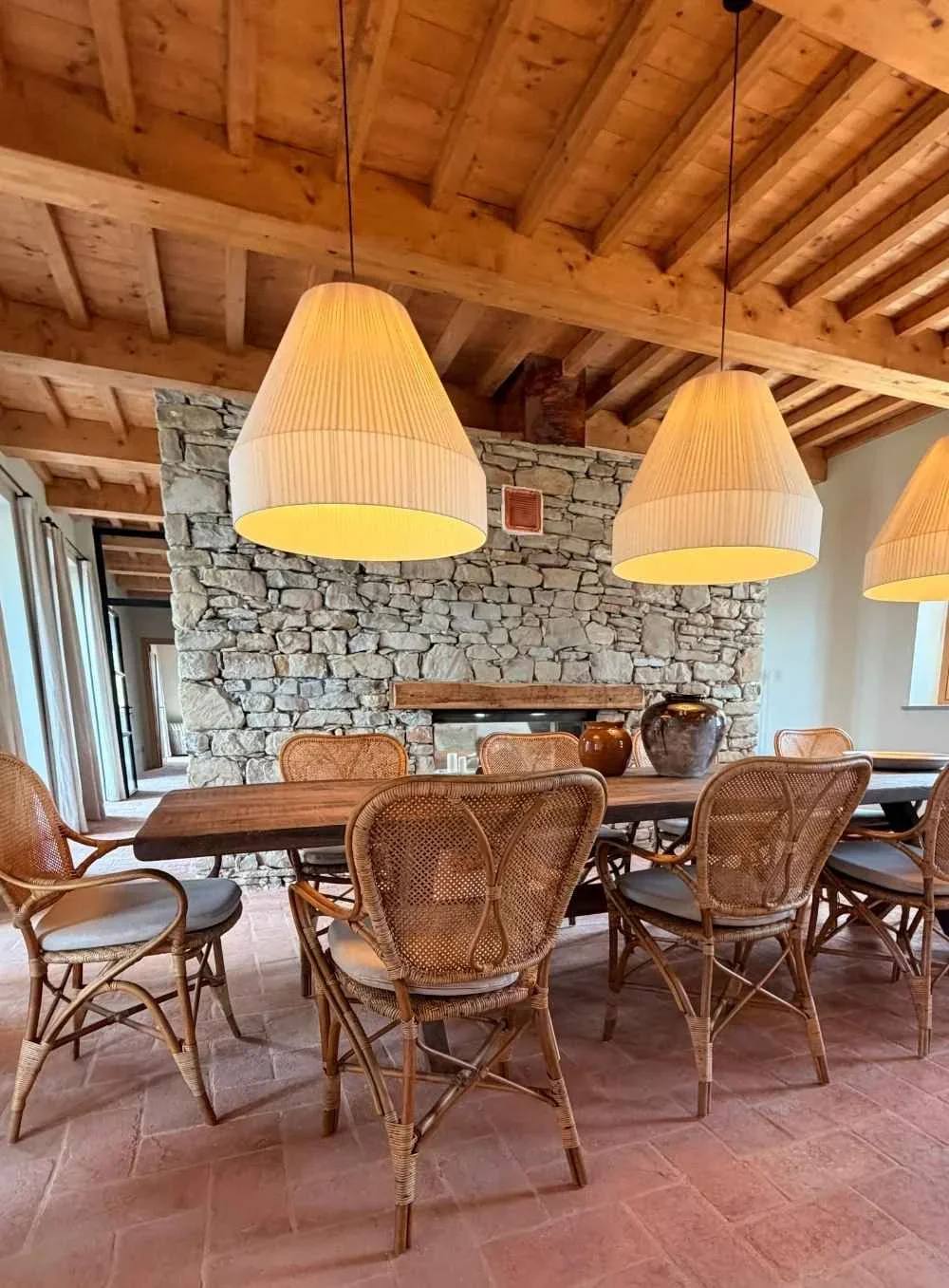 Dining room with a wooden table, wicker chairs with cushions, stone fireplace, wooden beamed ceiling, three hanging pendant lamps, and decorative vases on the table.