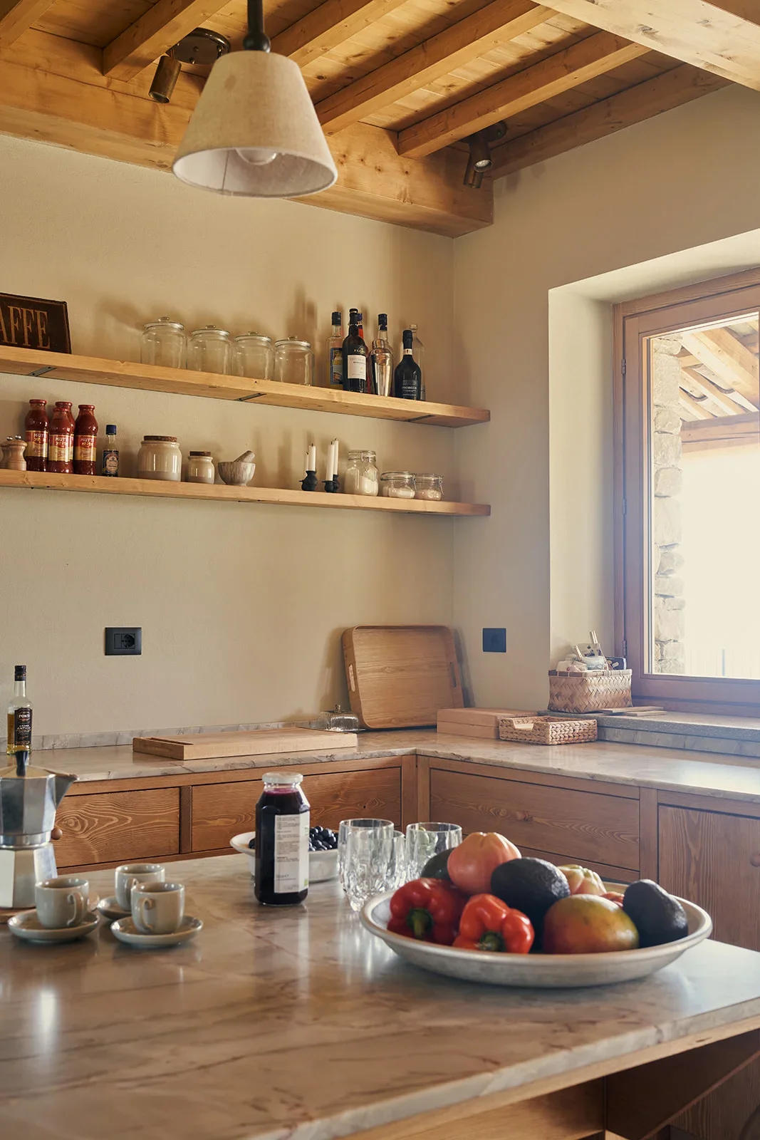 A cozy kitchen with wooden cabinets, shelves holding jars and bottles, a marble countertop with a fruit bowl, cups, and glasses, and a large window providing natural light.