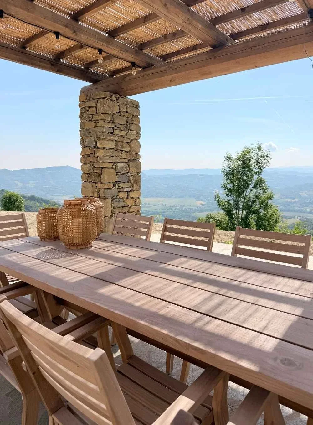 Outdoor dining area with a wooden table and chairs, decorative lanterns, stone pillar, wooden and bamboo roof, mountain view in the background.