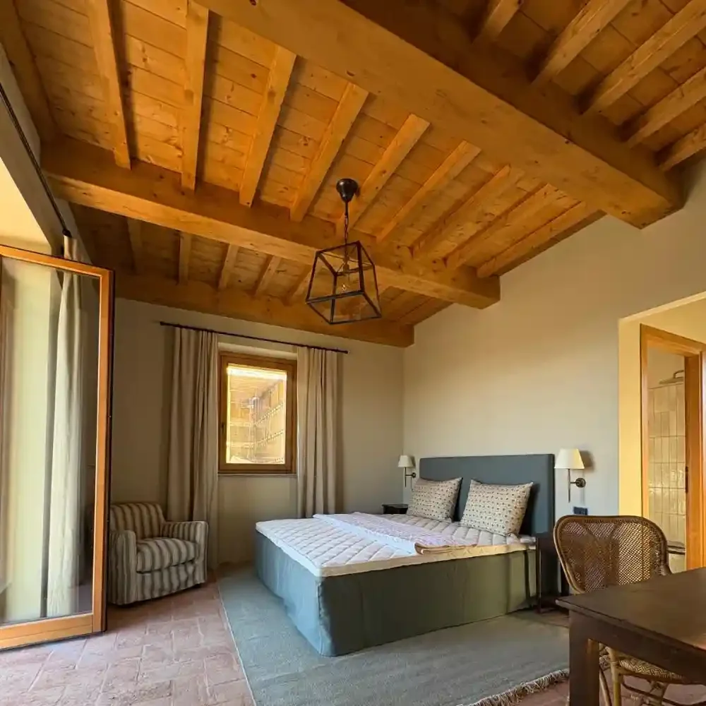A bedroom with a wooden ceiling and beige walls. There is a bed with pillows, a striped armchair in the corner, a window with curtains, and a wooden door. A hanging light fixture is attached to the ceiling.