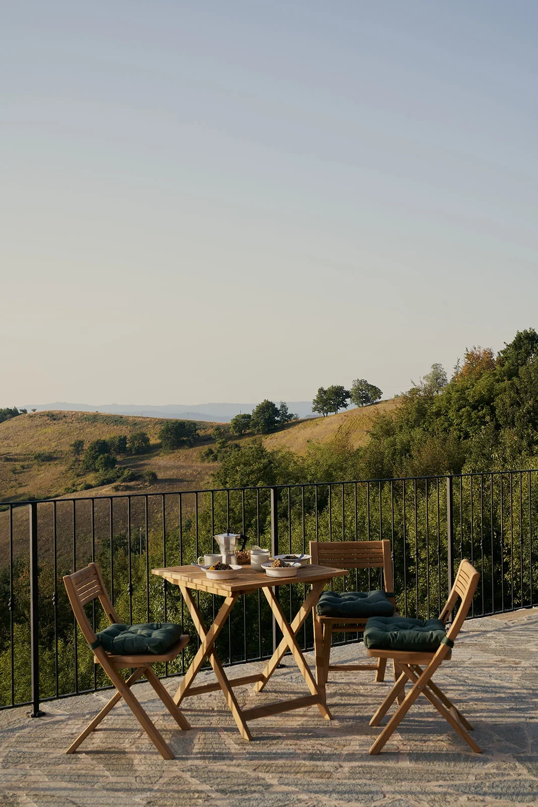 Outdoor balcony with a wooden table and four chairs, set with breakfast items, overlooking rolling hills and trees at sunrise or sunset.
