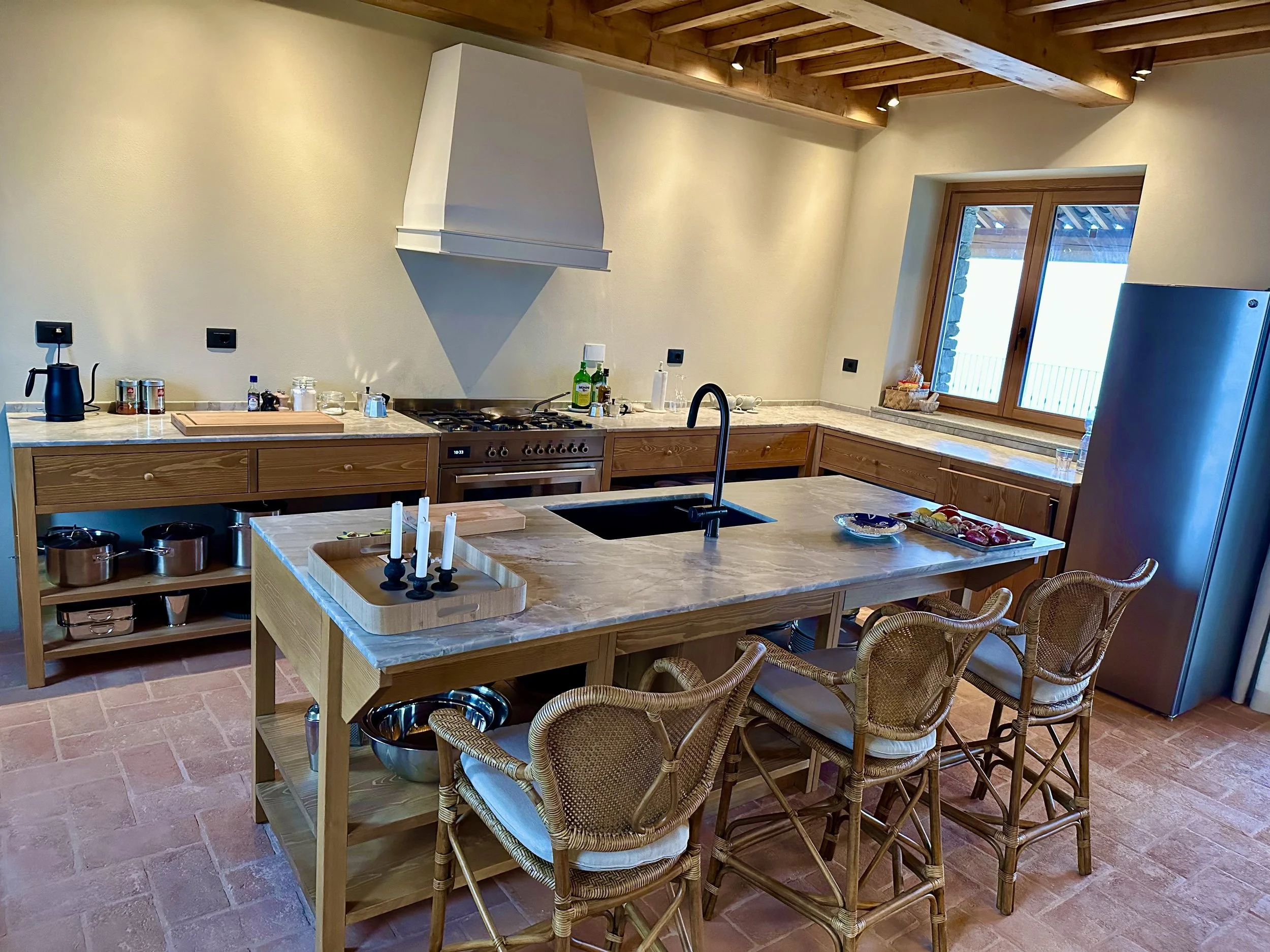 Kitchen with wooden cabinets, a central island with a marble countertop, four wicker chairs, stove with a range hood, window above the counter, and various small kitchen items and appliances.