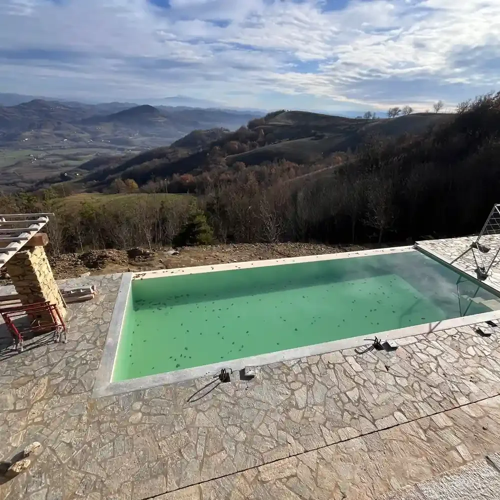 A swimming pool with green water on a stone patio overlooking rolling hills and a cloudy sky in the distance.