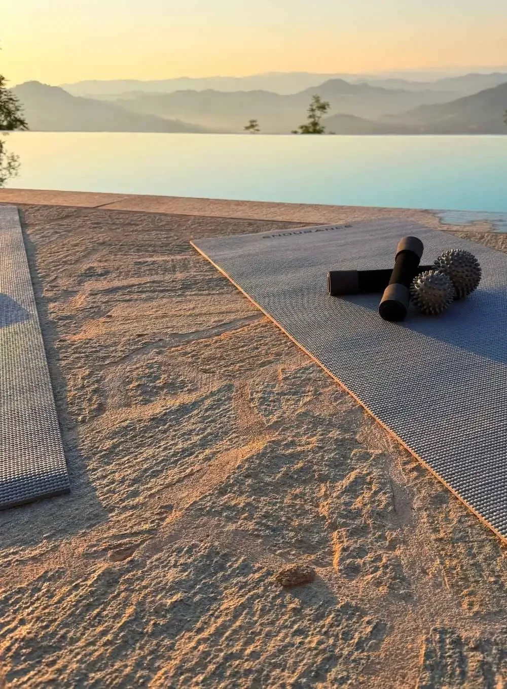 Yoga mats and massage balls on a stone surface with a pool and mountain view at sunset.
