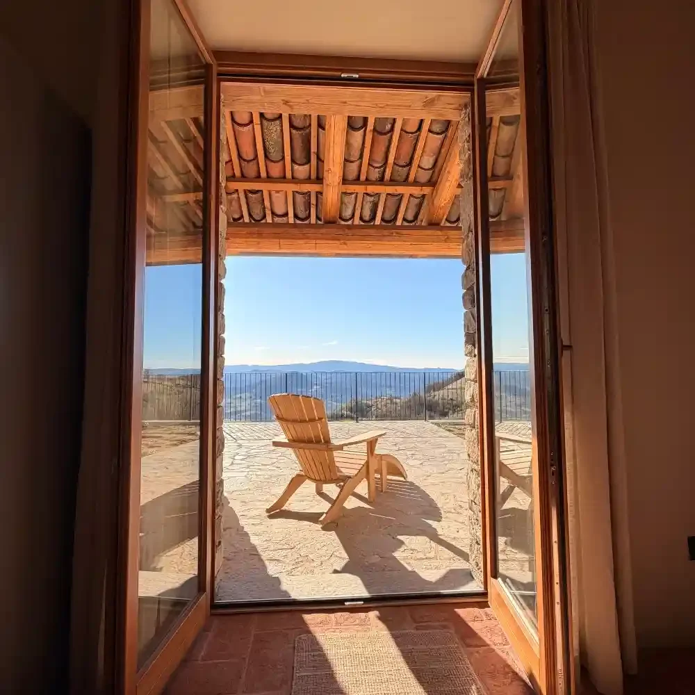 Balcony with a wooden Adirondack chair overlooking mountains and a clear blue sky, framed by open glass doors.