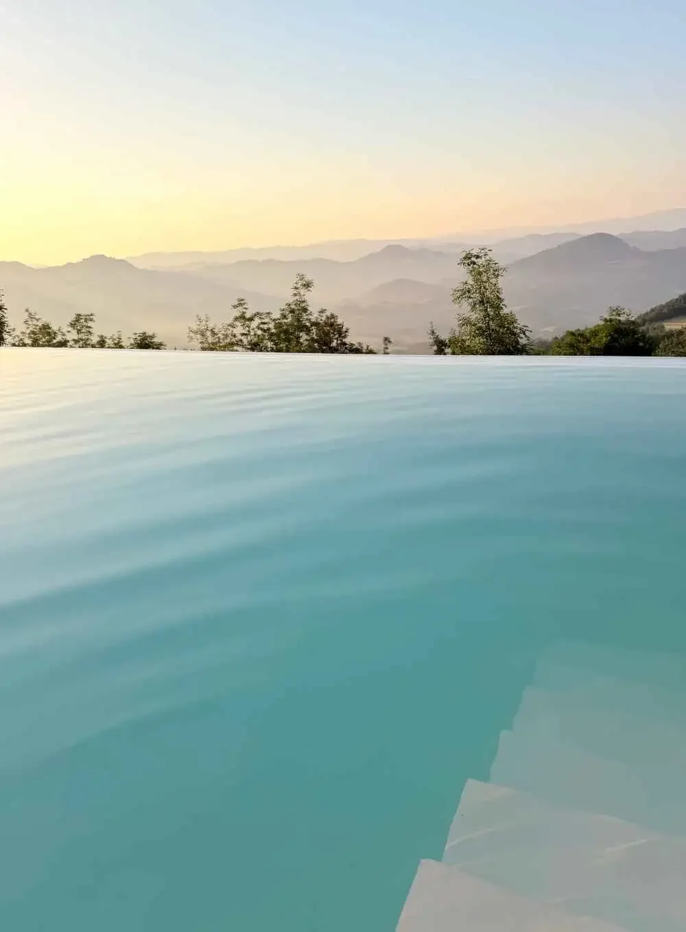 Photo of a swimming pool with water in the foreground, overlooking a landscape of distant mountains and trees during sunset or sunrise.