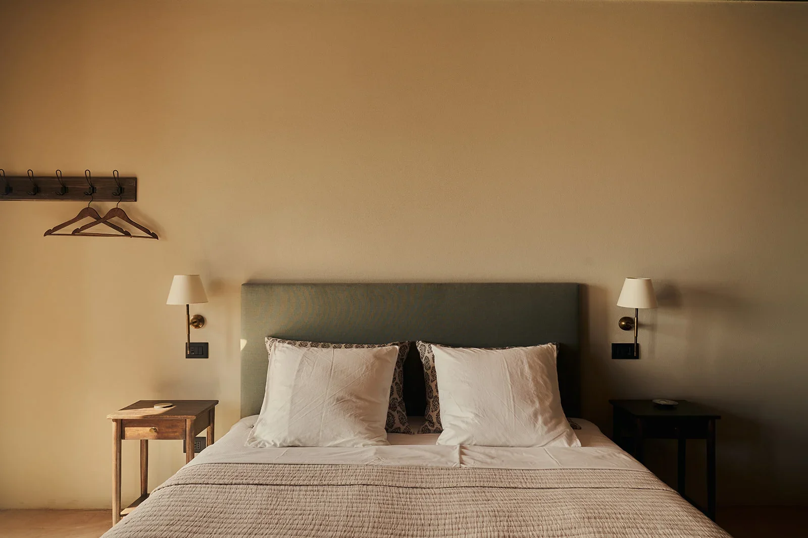 A neatly made bed with a beige quilt, two large white pillows, and two decorative pillows with a leopard print. The bed has a teal headboard. On each side of the bed, there are black nightstands with small lamps attached to the wall above each. To th