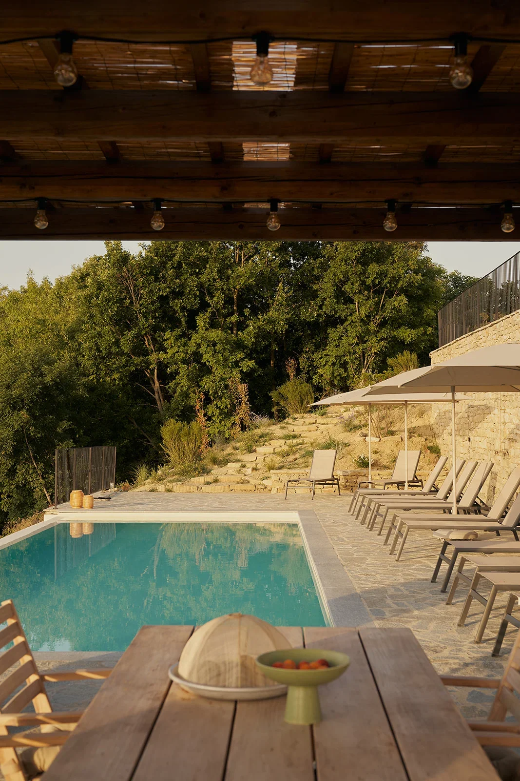 A backyard pool area with lounge chairs, umbrellas, and a wooden dining table with a bowl of fruit, view from under a wooden pergola, surrounded by trees and stone steps.