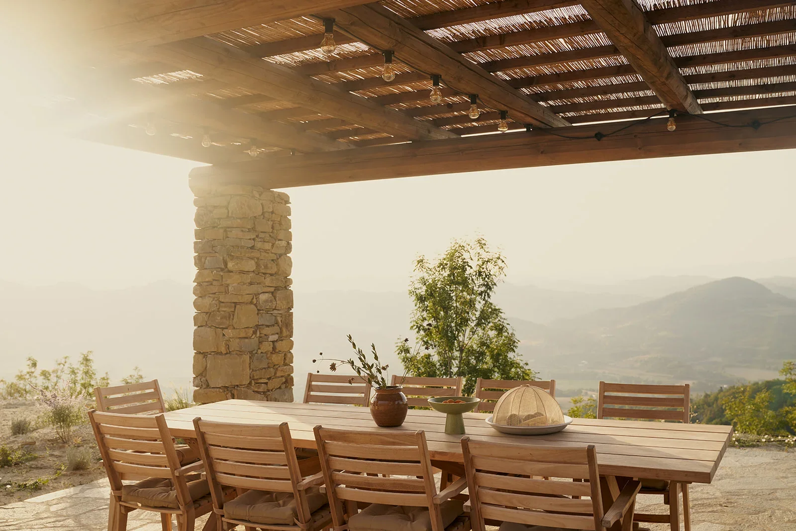 Outdoor dining area with a long wooden table and eight matching chairs, set under a wooden pergola with a stone pillar, overlooking a mountainous landscape during sunset.