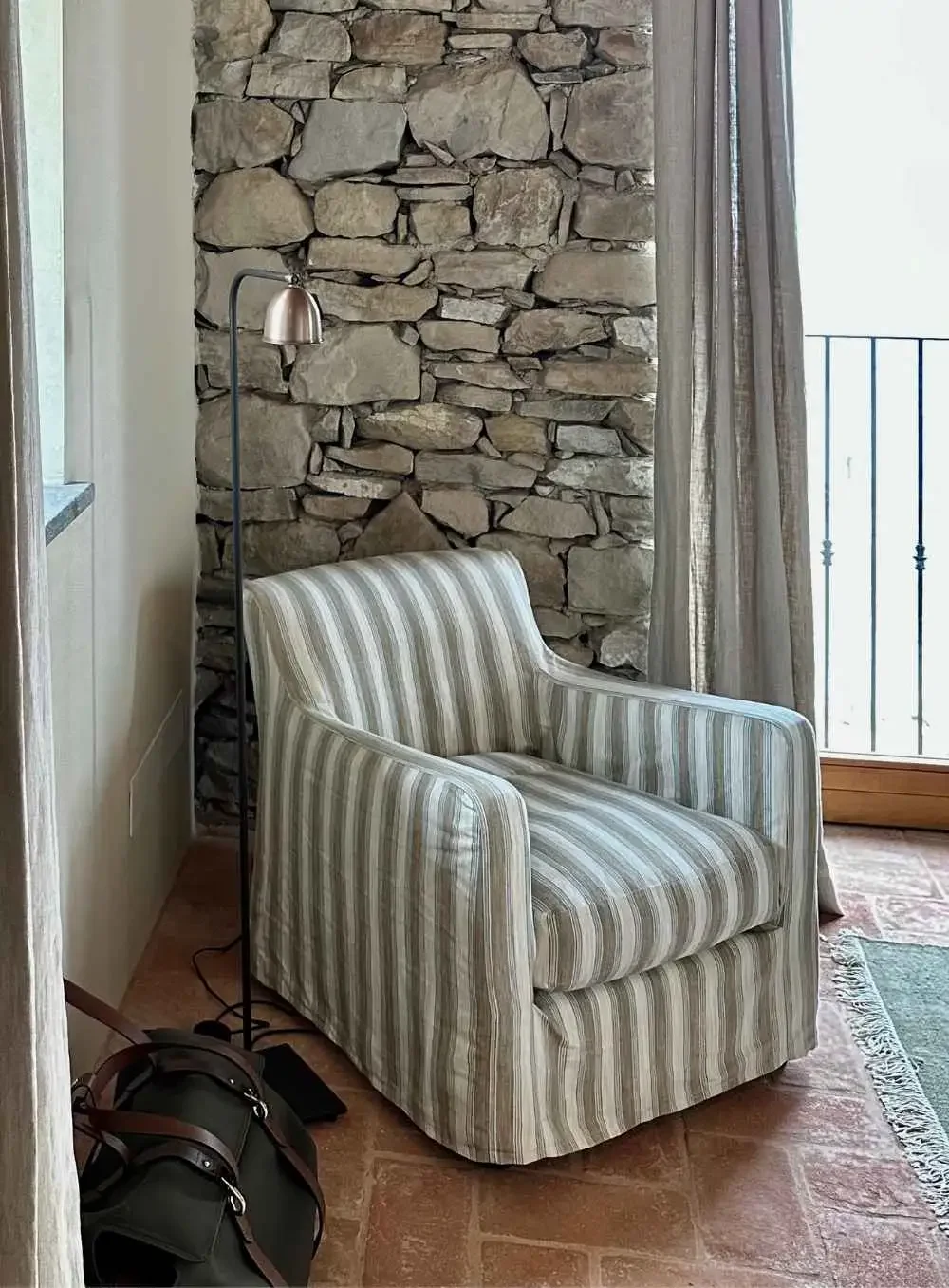 A striped armchair next to a floor lamp with a stone wall behind it, next to a window with curtains and a balcony railing outside.