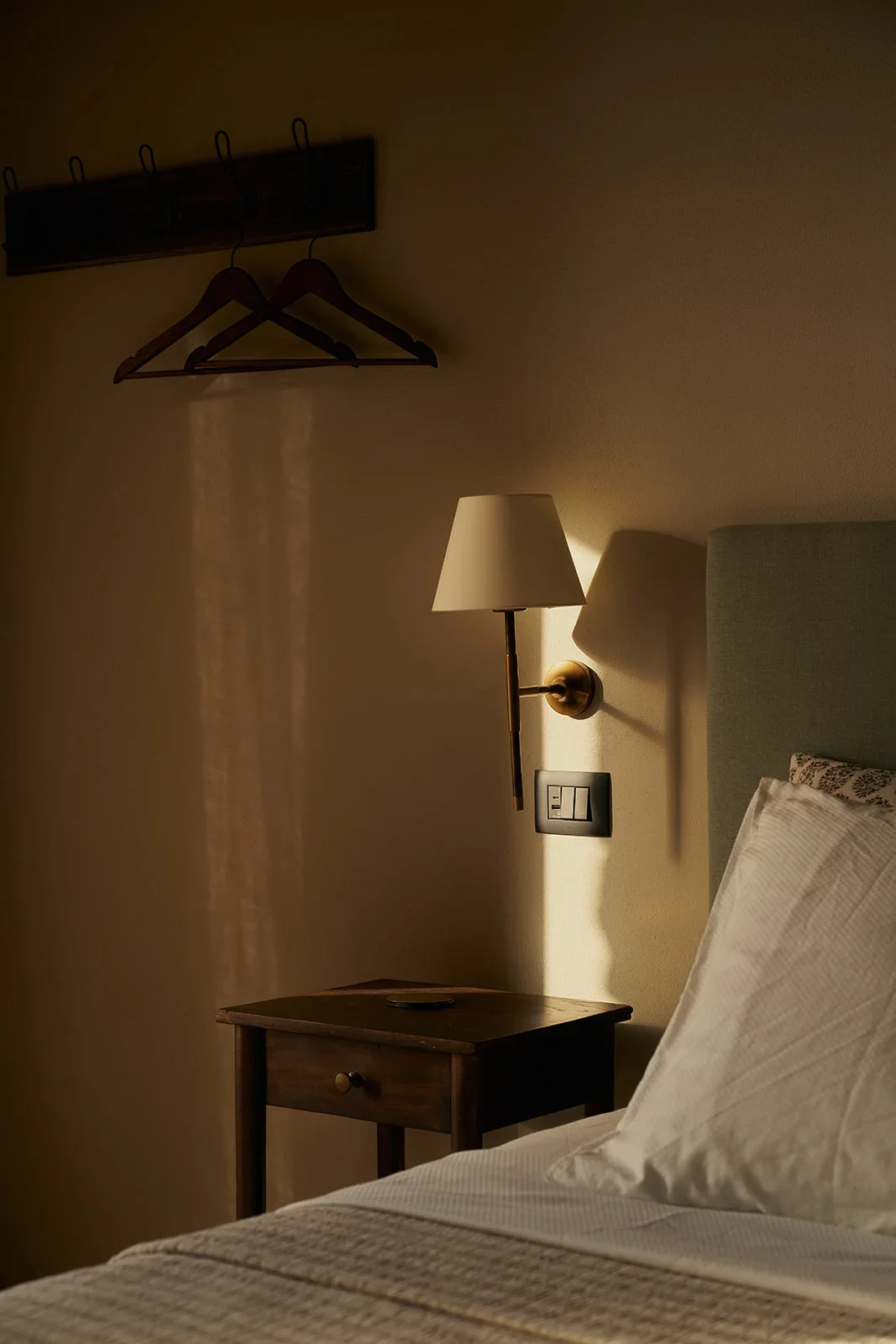 A hotel room with a lit wall-mounted lamp, a wooden nightstand, a bed with white linens, a headboard, and two empty coat hangers on a wall hook.