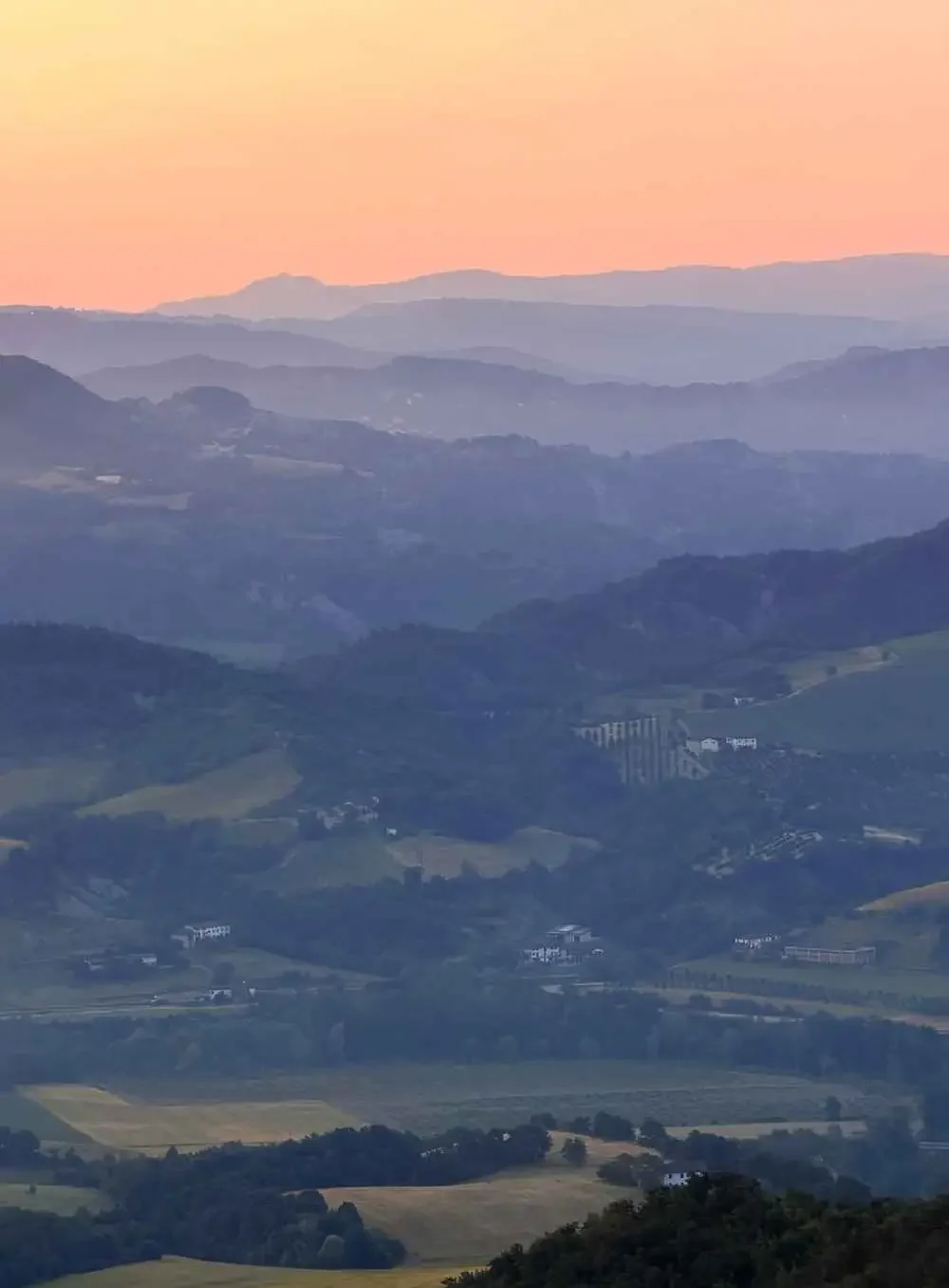A landscape of rolling hills and mountains at sunset with a pink-orange sky, showing multiple layers of mountains fading into the distance.