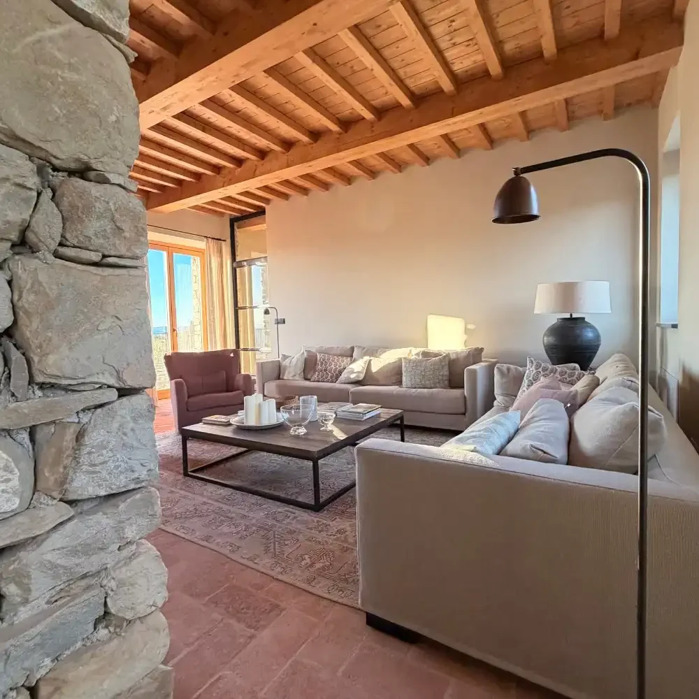 Living room with stone wall, wooden ceiling, beige sofas, an armchair, a coffee table with candles and books, a large floor lamp, and a large window with wooden framing.