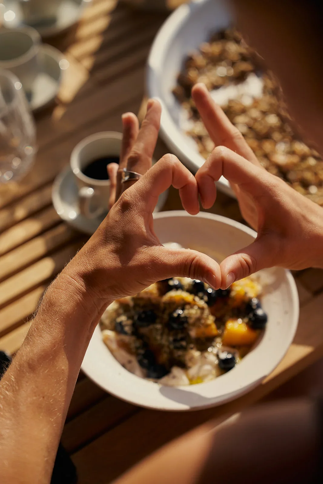 Person making a heart shape with hands over a bowl of fruit and desserts on a wooden table.