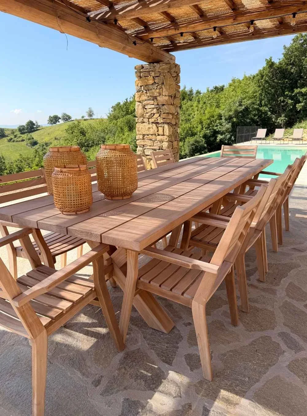 Outdoor patio with a large wooden dining table, wooden chairs, decorative wicker lanterns, a stone pillar, a wooden pergola, a swimming pool, and green trees in the background on a sunny day.