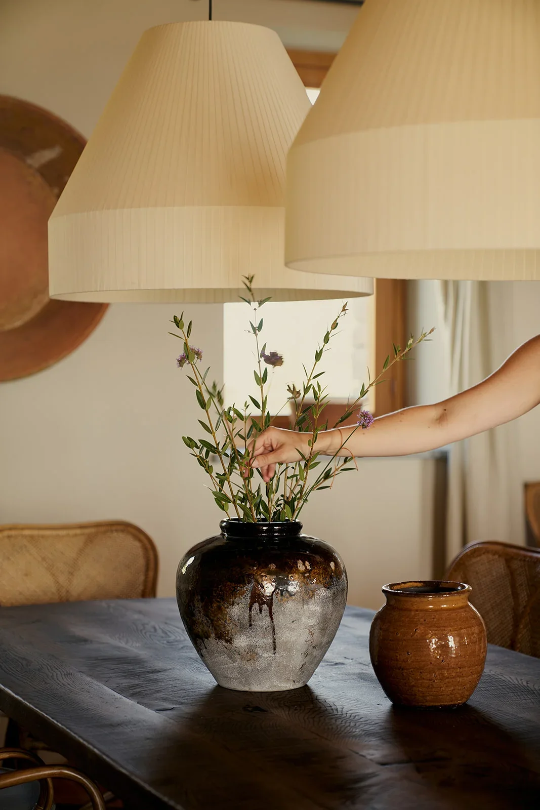 A hand arranging purple flowers and green foliage in a dark ceramic vase on a wooden table, with two large beige lamps hanging overhead and wooden chairs in the background.