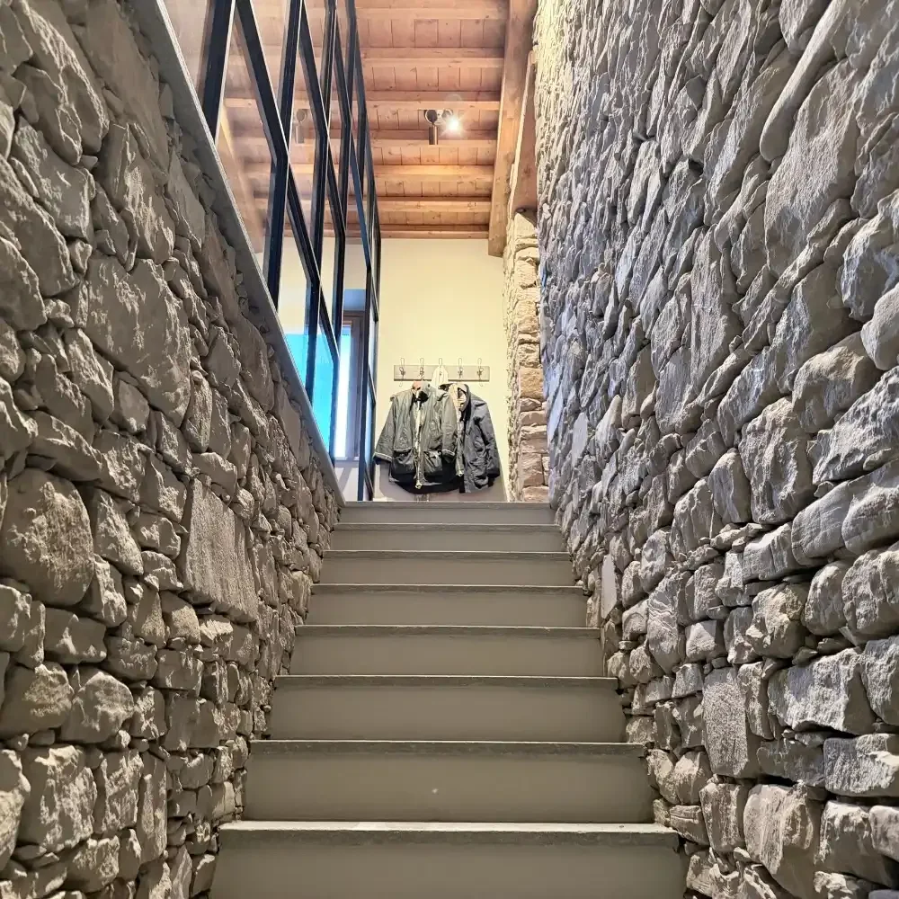 Stone walls on either side of a stairway with gray steps and a wooden ceiling. At the top, there is a clothing rack with jackets hanging on it.