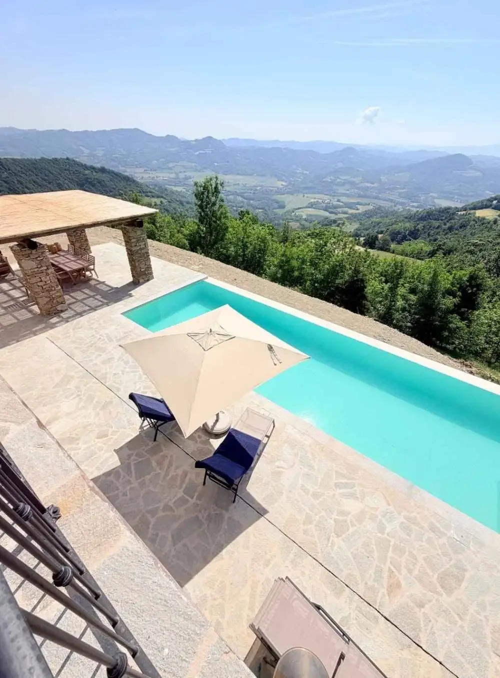 View of a swimming pool on a stone patio overlooking green hills and mountains under a blue sky with some clouds.