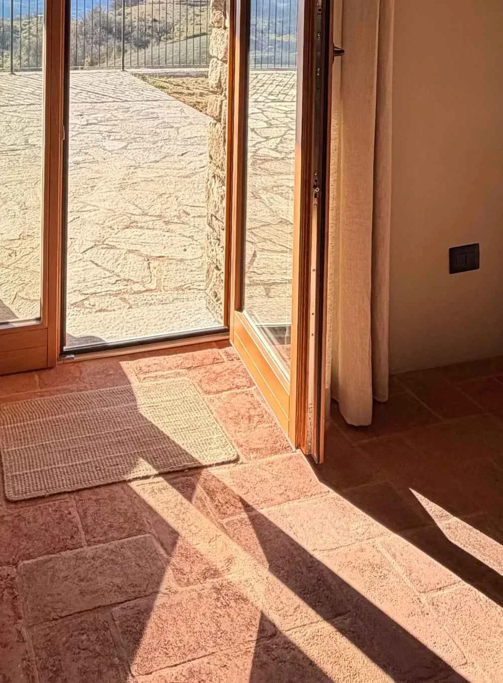 Sunlight casts shadows inside a room through an open glass door leading outside to a sunlit stone patio. The door has a wooden frame, and a small rug is placed on the terra cotta tiled floor near the door.