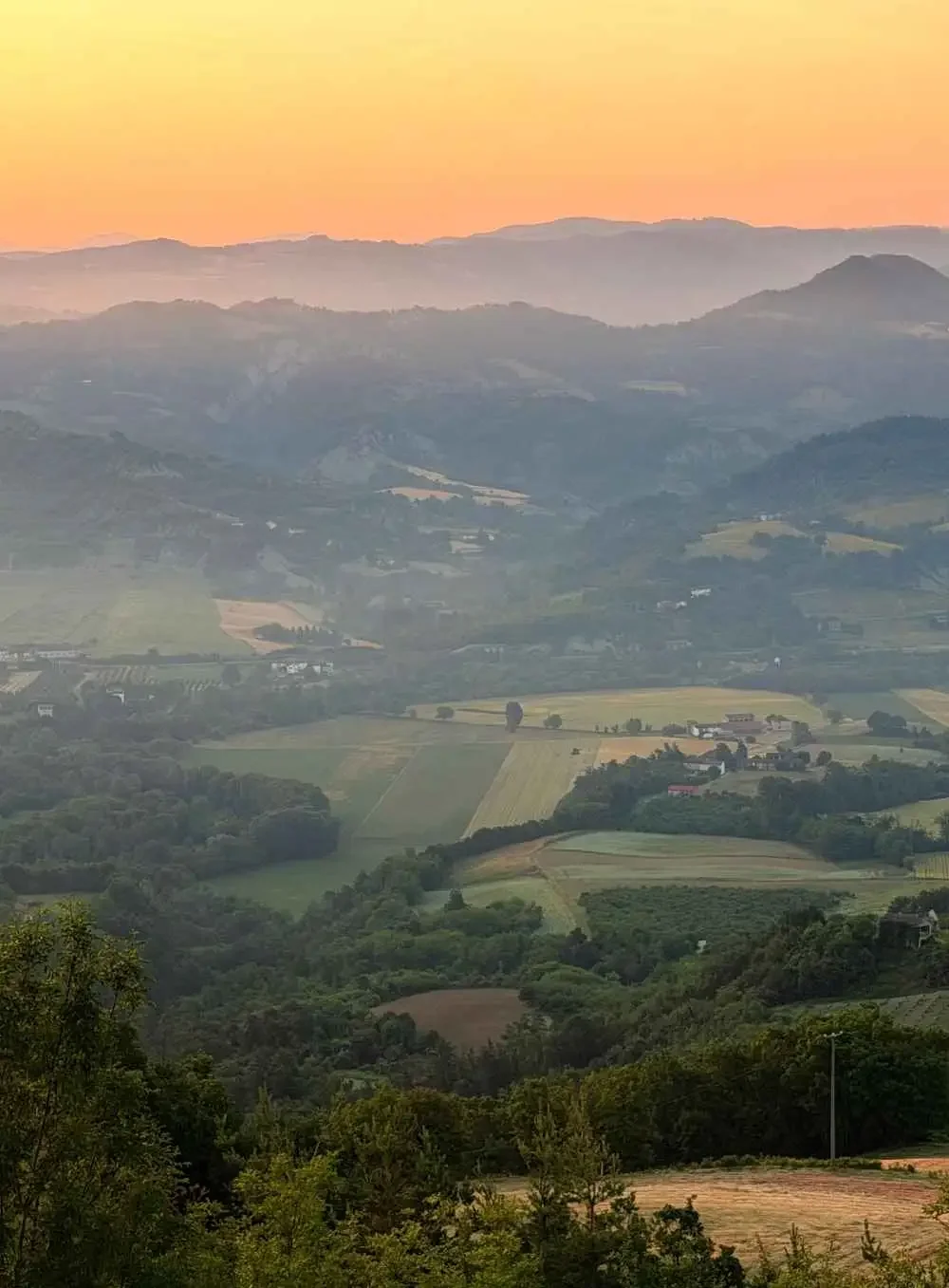 A scenic landscape of rolling hills and valleys with patches of farmland, under a pastel-colored sky at sunset or sunrise.