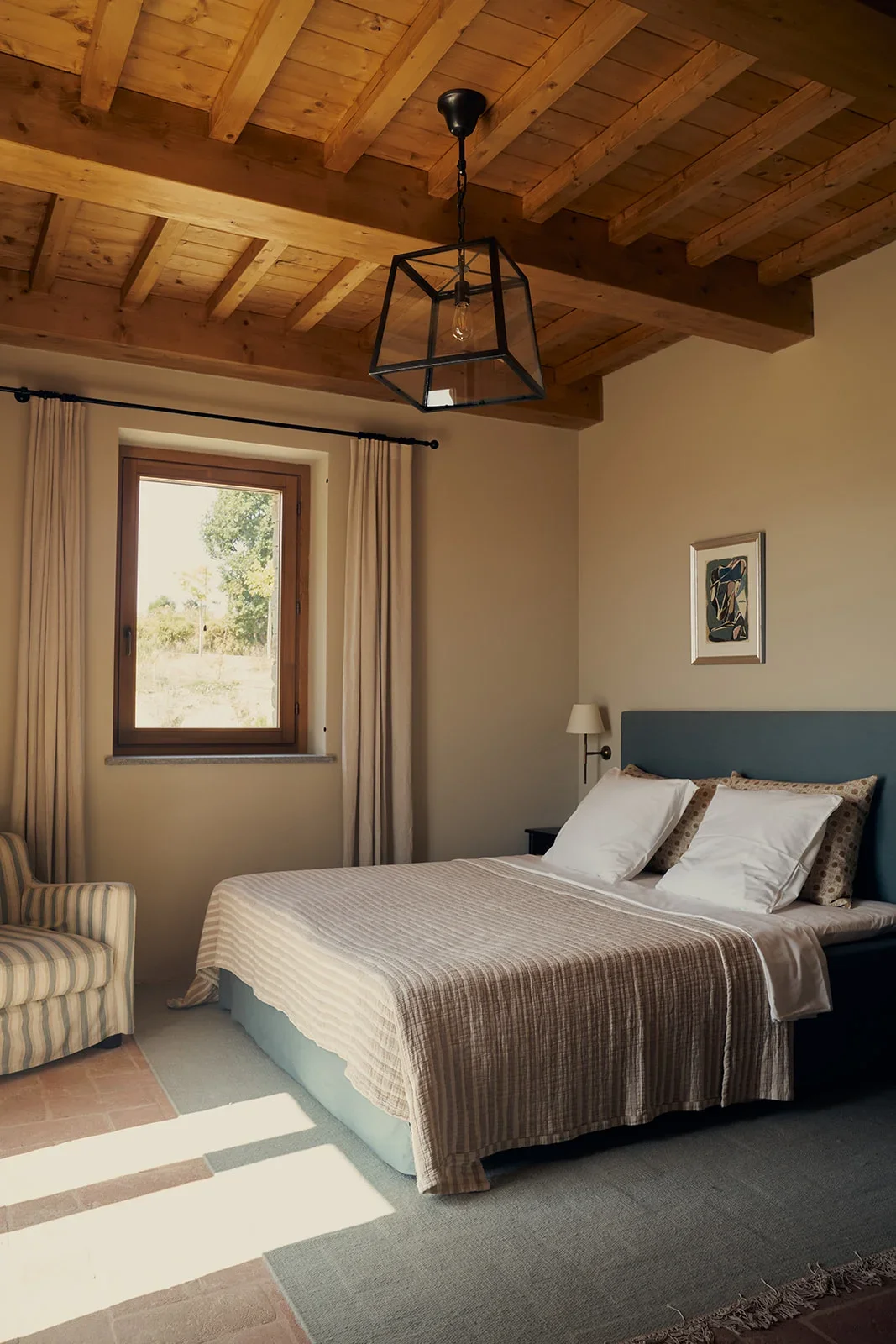 A cozy bedroom with a wooden ceiling, a window with curtains, a bed with pillows and a blanket, a bedside lamp, a framed picture on the wall, and an armchair.