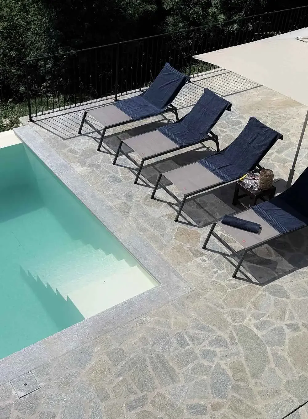 A swimming pool with a stone deck, three lounge chairs with dark blue towels, small side table with a lantern and drinks, and a white patio umbrella, with greenery and trees in the background.