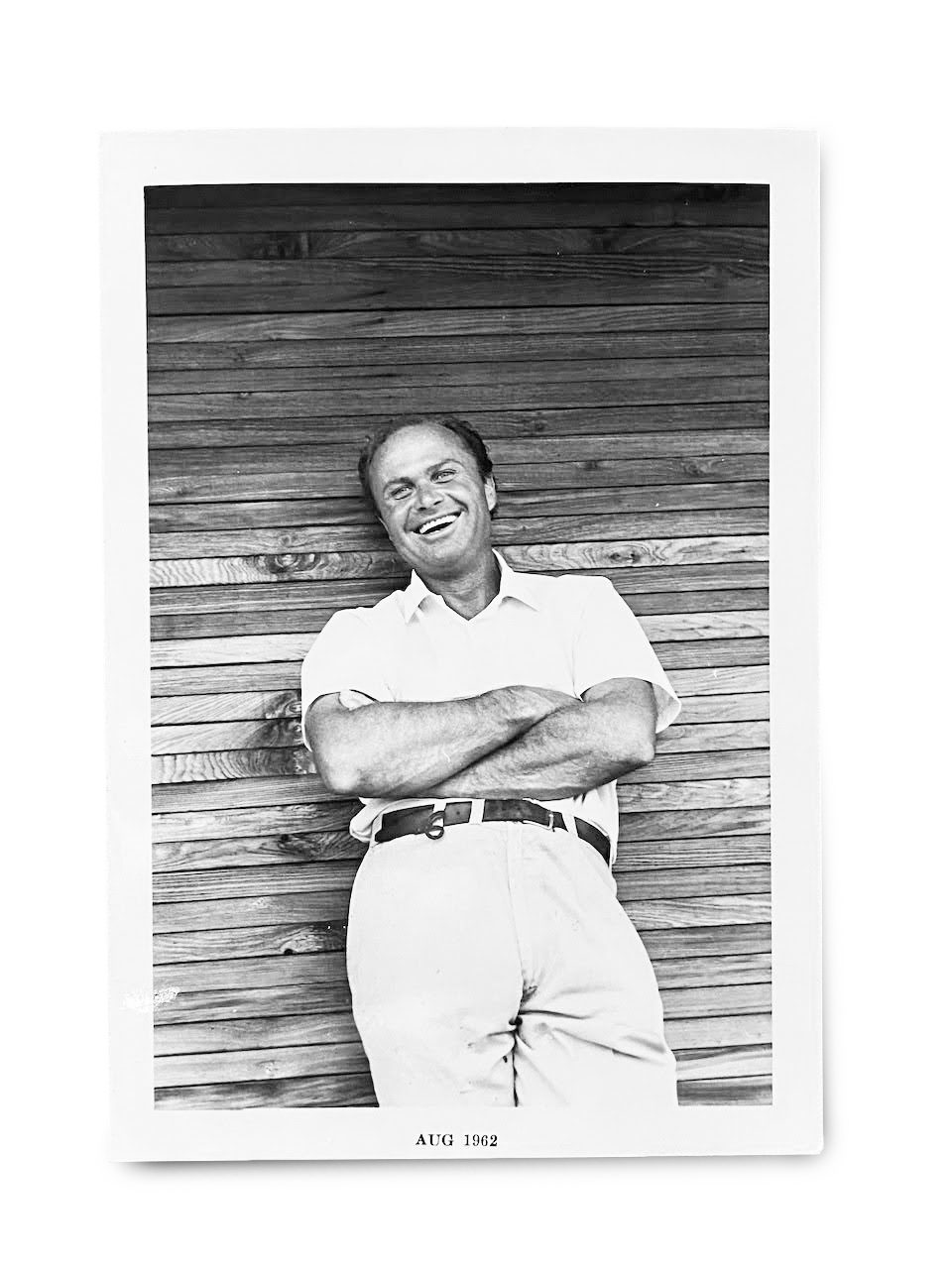 A black and white photo of a smiling man (Emile Norman) with crossed arms, standing in front of a wooden wall. The photo is labeled "AUG 1962."