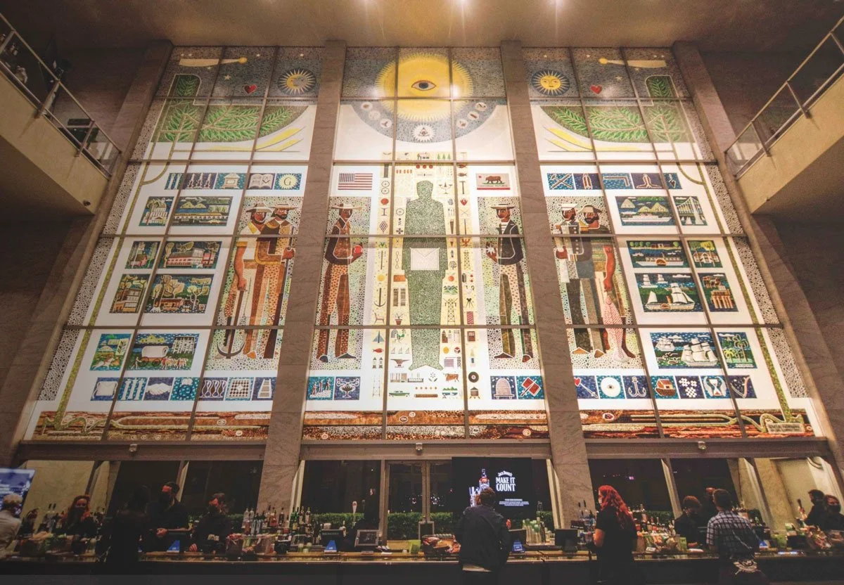 A large, colorful mosaic window depicting people, symbols, and imagery representing American culture and history, located on the wall behind a bar with people gathered around.