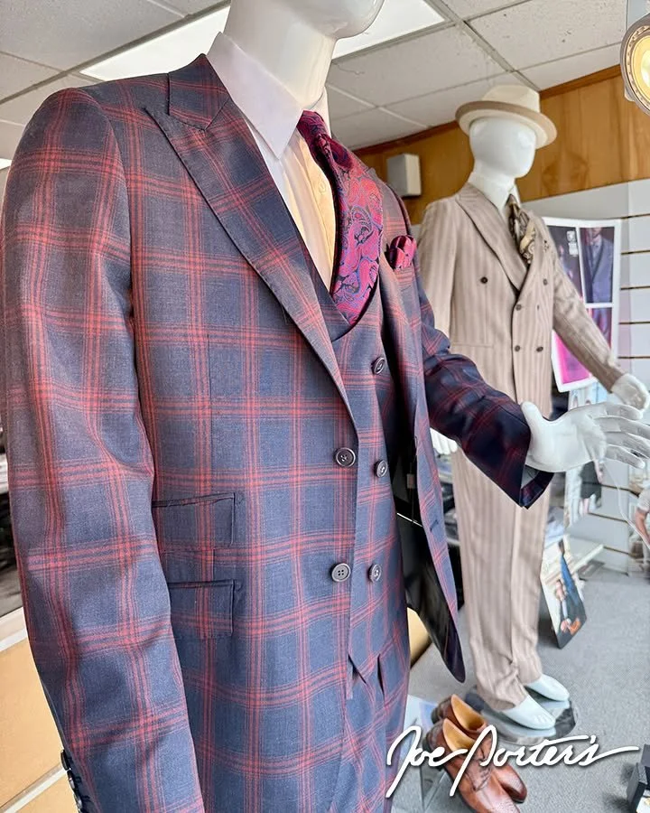 Mannequins display a navy and red plaid three-piece suit with a pink paisley tie and pocket square, alongside a beige pinstripe double-breasted suit in a menswear store.