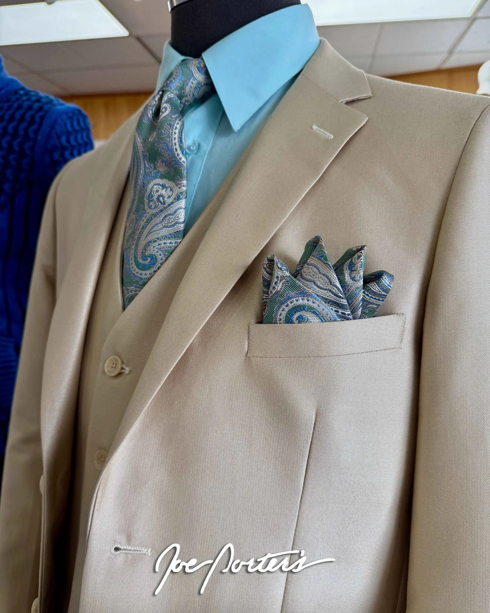 Close-up of a beige men's suit with a light blue dress shirt, paisley tie, and matching pocket square.