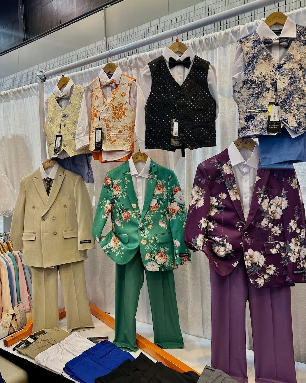 Retail display of colorful men's formal suits, floral blazers, dress shirts, and patterned vests on mannequins and hangers