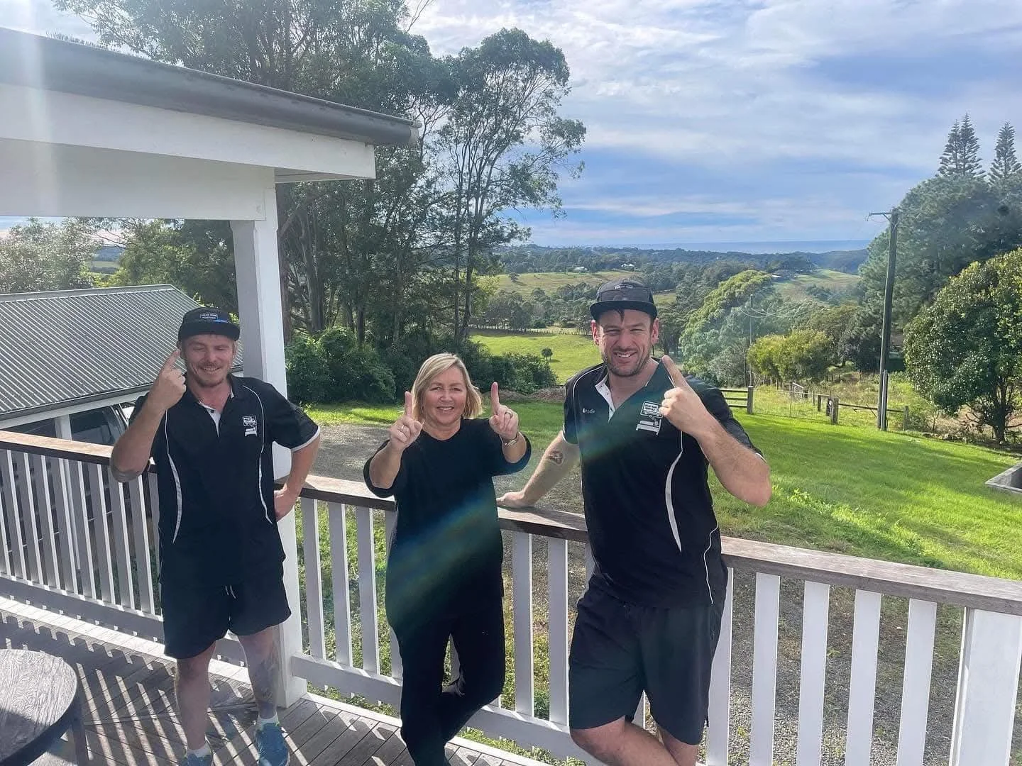 Three people standing on a wooden deck outdoors, smiling and making gestures, with green rolling hills and trees in the background under a partly cloudy sky.