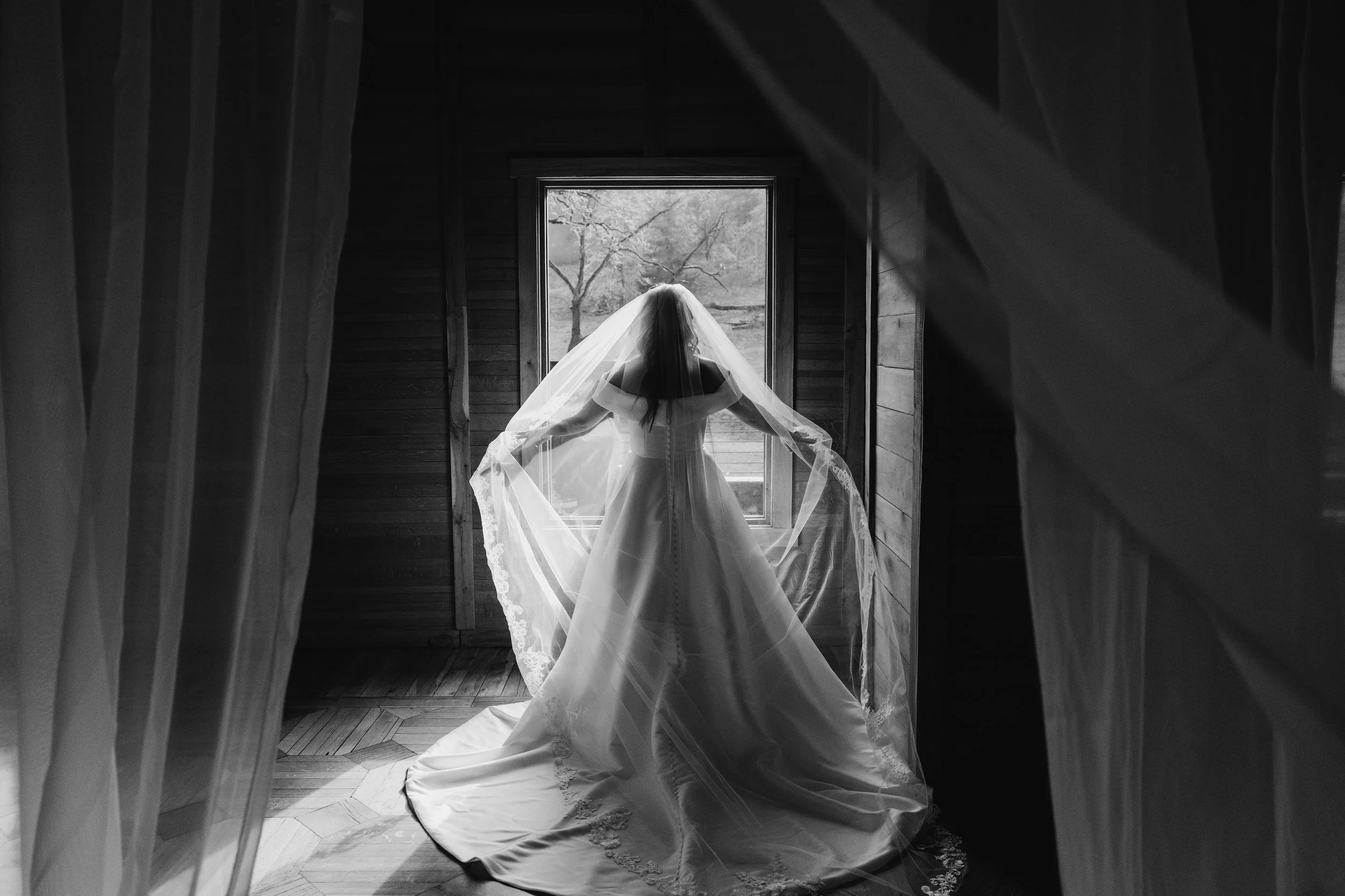 Black and white portrait of bride standing in front of window in her Stella York wedding gown and cathedral veil