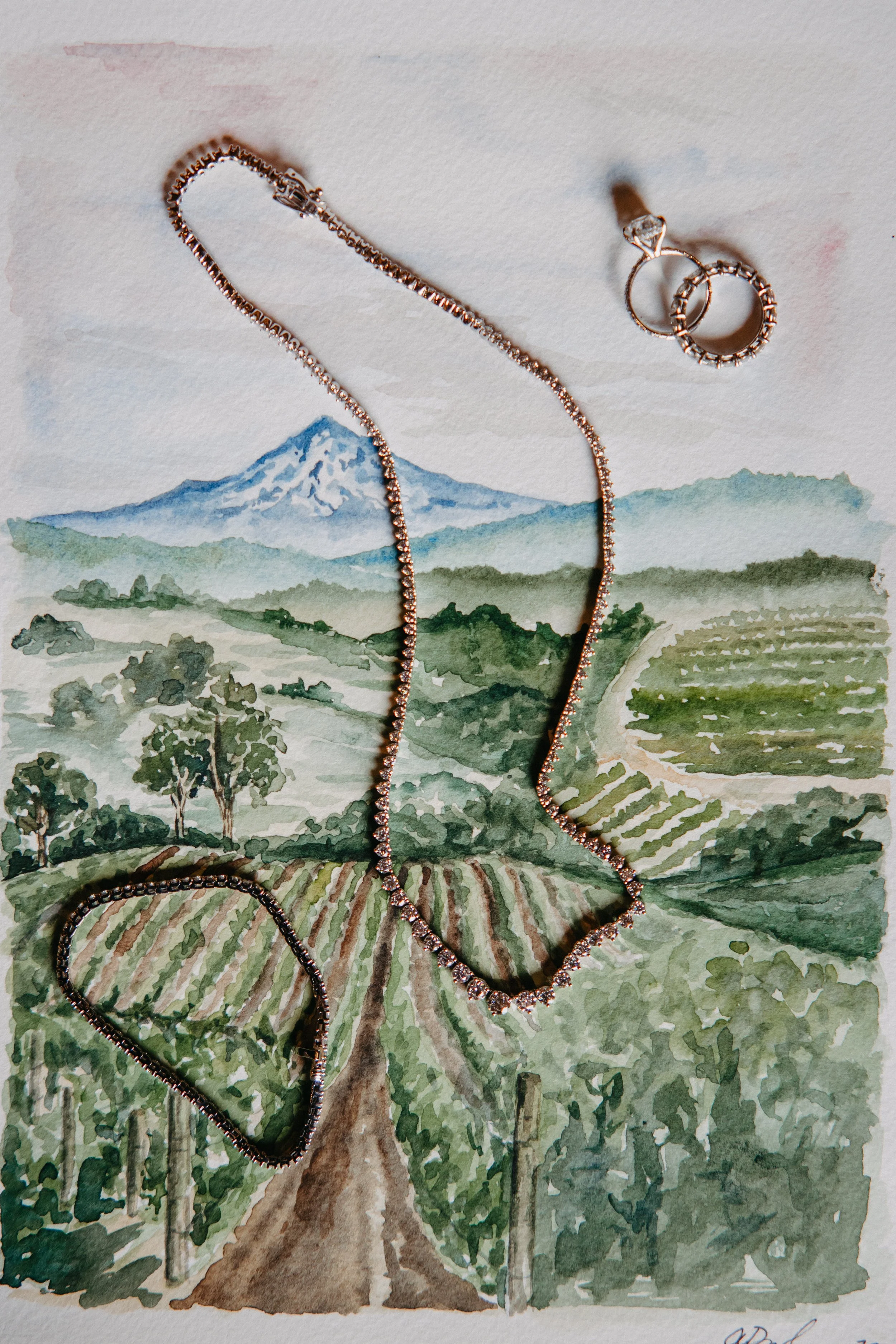 Watercolor painting of Mt.Hood with vineyards in front and wedding jewelry placed on top of painting