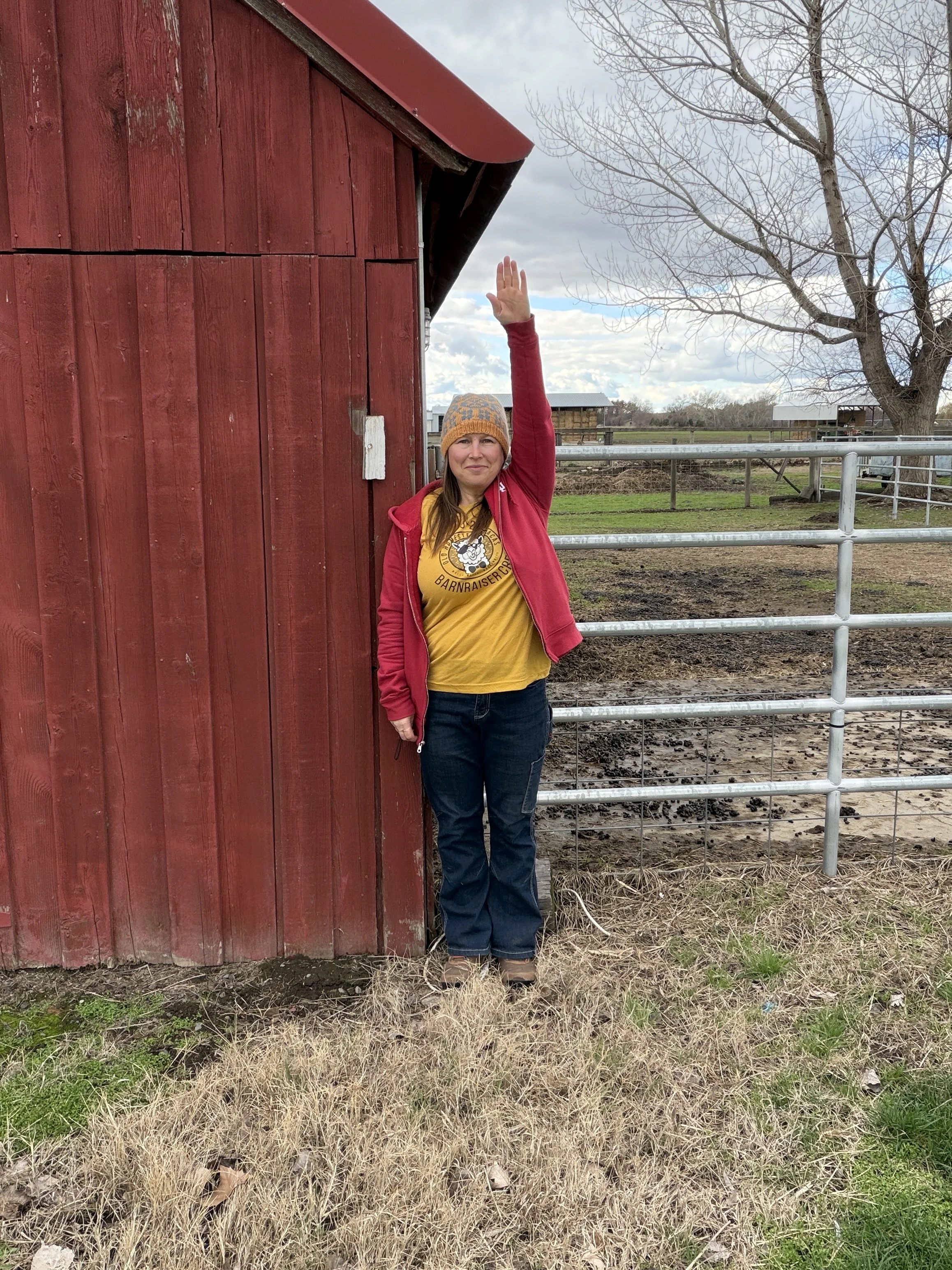 A woman standing outdoors next to a red wooden barn, raising her right hand. She is wearing a yellow shirt with a graphic and text, a red jacket, and a harnessed beanie hat, with a partly cloudy sky and leafless tree in the background.