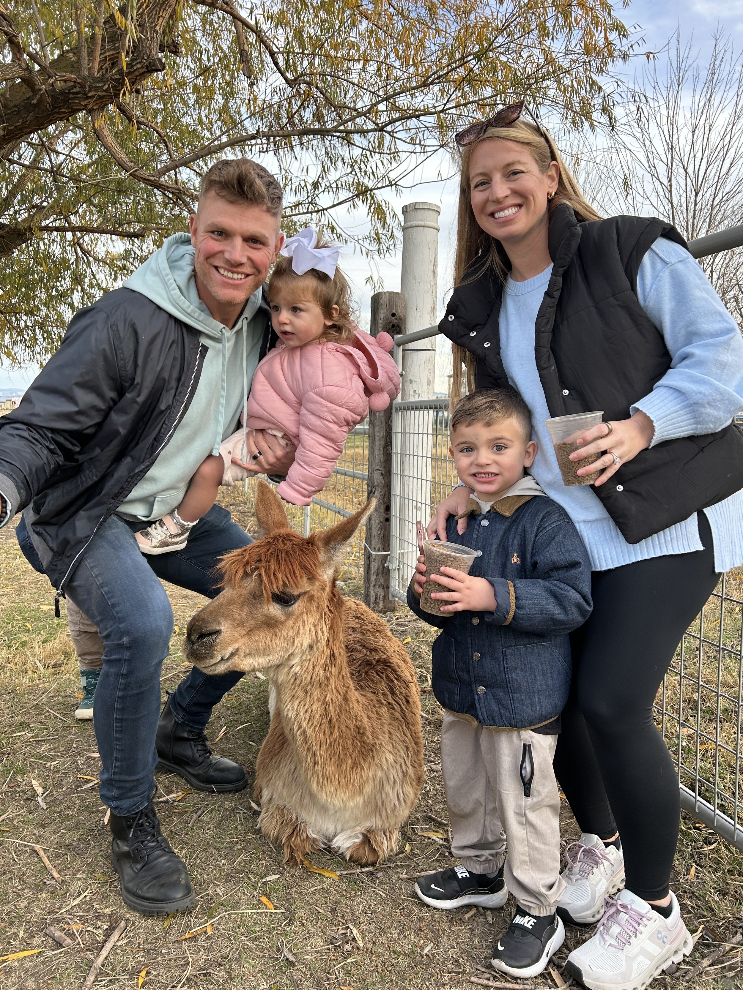A family of four visiting a farm or petting zoo, standing next to a brown alpaca, holding cups of food, with trees and a fenced enclosure in the background under partly cloudy skies.