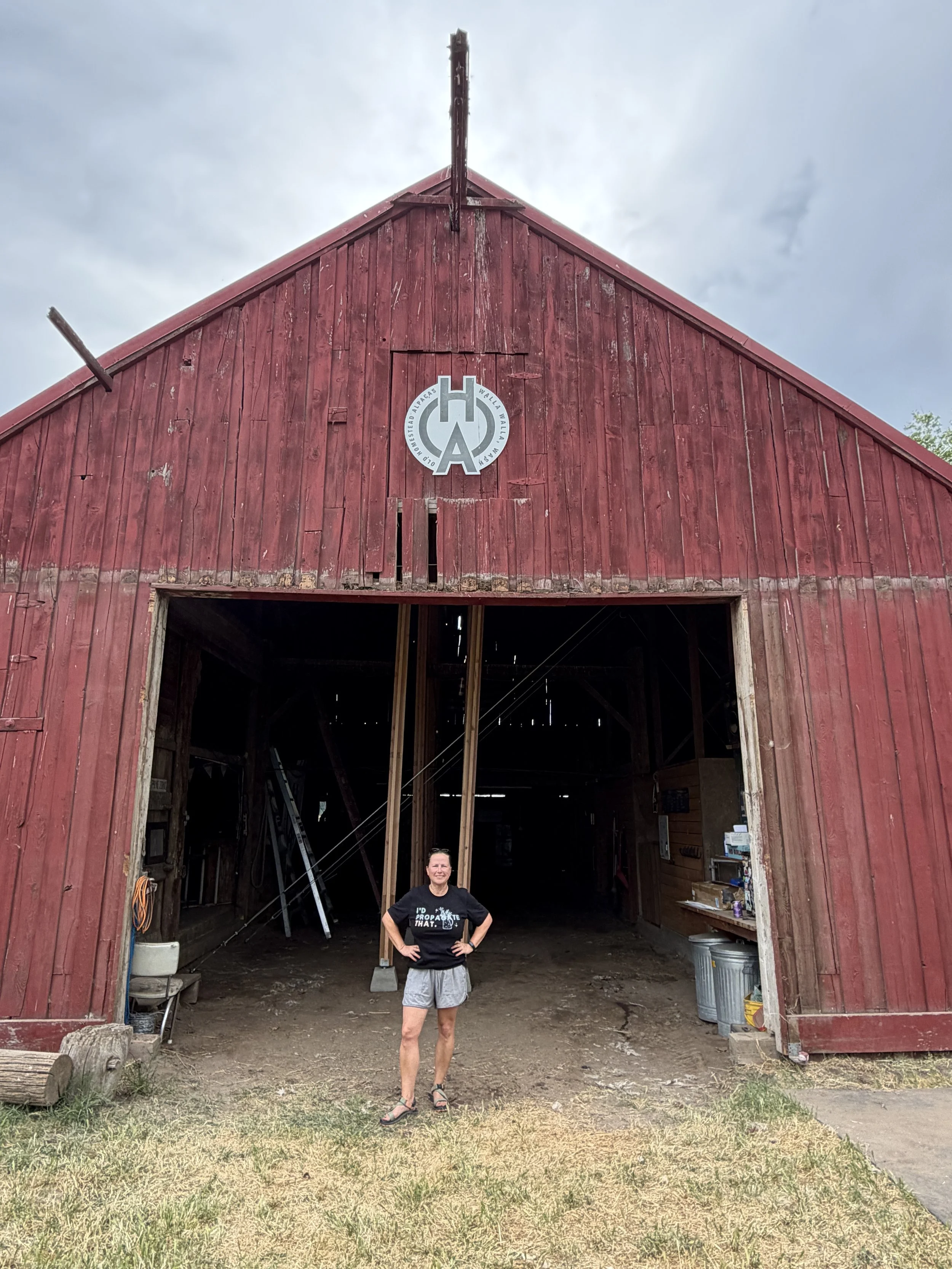 A woman standing in front of an old red barn under a cloudy sky. She is wearing a black t-shirt, shorts, and sandals, smiling with her arms on her hips.