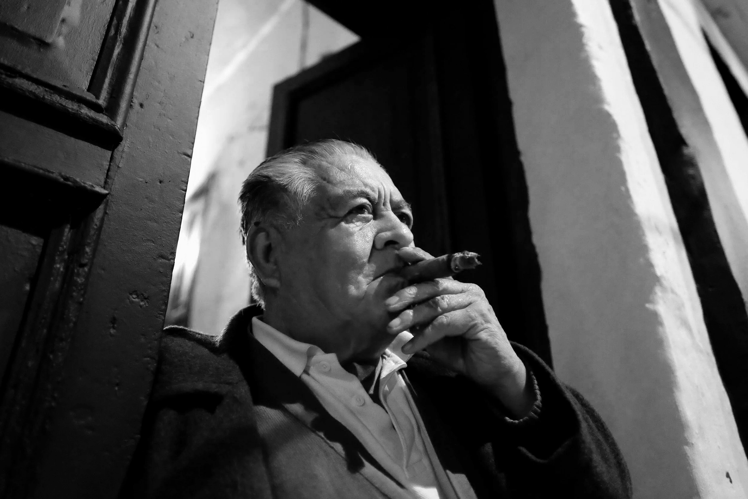 An elderly man with slicked-back hair, smoking a cigar, sitting on a staircase in a building with black and white interior, wearing a collared shirt and dark coat.