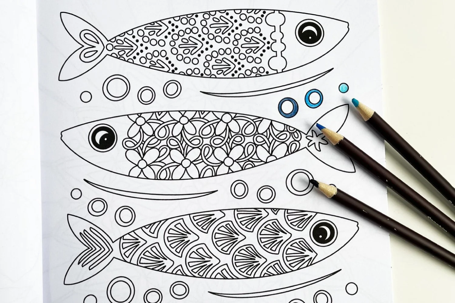 Close up photo of a colouring page featuring patterned sardine fish. The line art has a small amount of colouring in and pencils are beside the book.