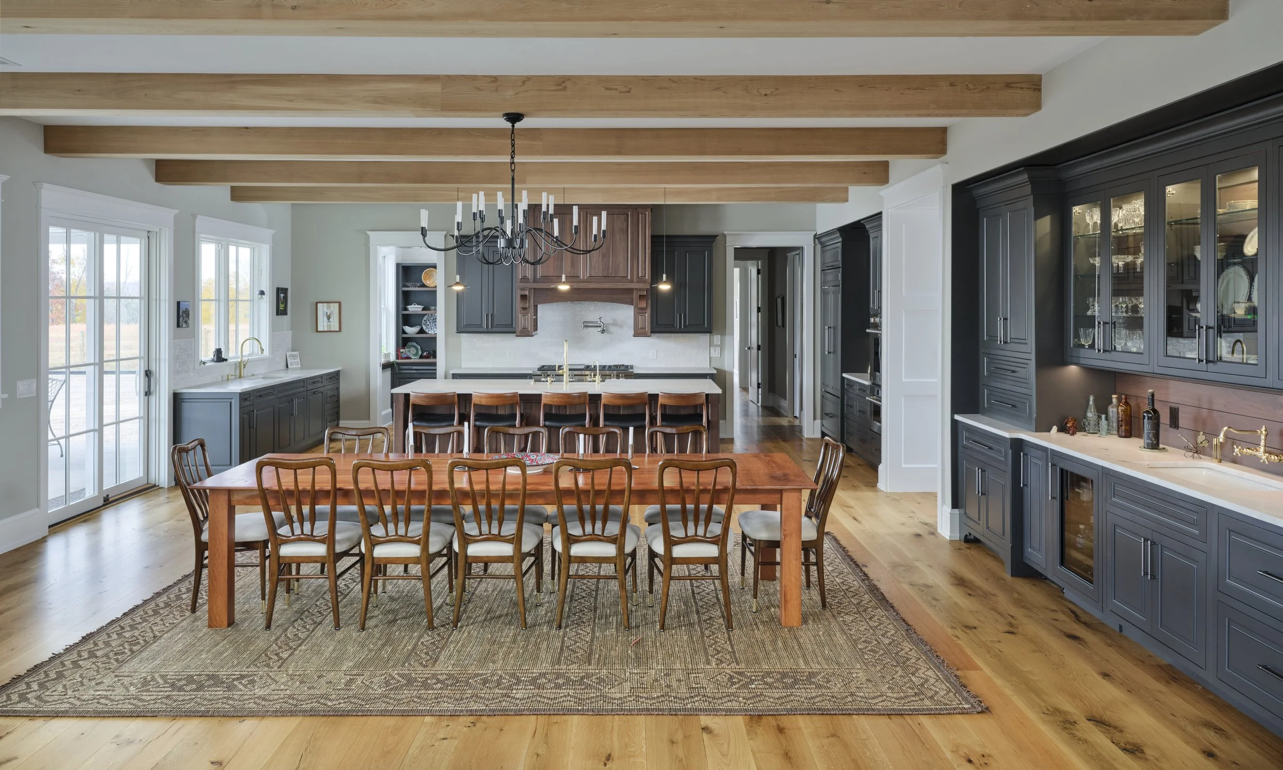 Open-concept kitchen and dining area with natural wood beams on the ceiling, black cabinetry, a large wooden dining table with ten chairs, and a kitchen island with barstools. There's a sliding glass door, multiple windows, and built-in shelves with 