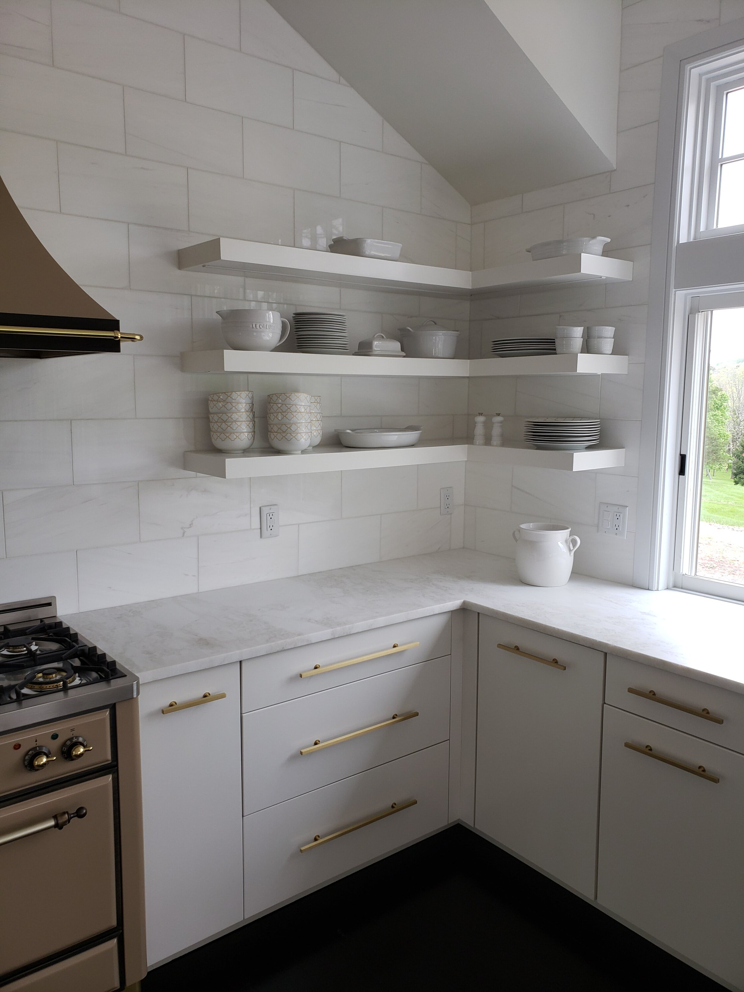 White kitchen with open shelves holding white dishes, bowls, and cups; marble countertops; a window with outside greenery; a white ceramic pot on the counter.